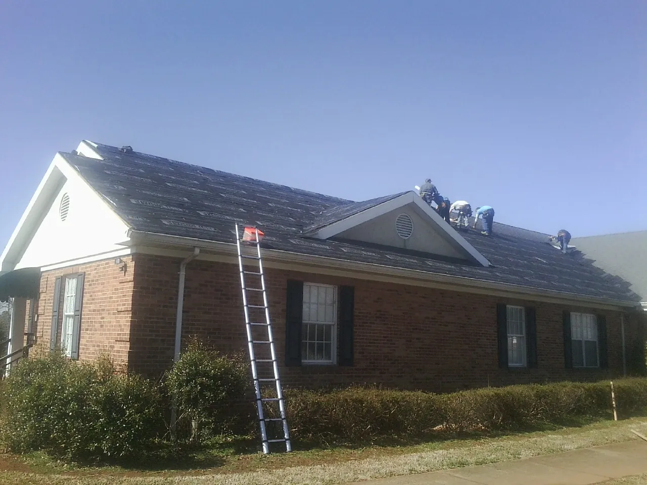 Crew tearing shingles off a brick ranch roof with a ladder set — Emergency Roof Repair work by our crew in Calera