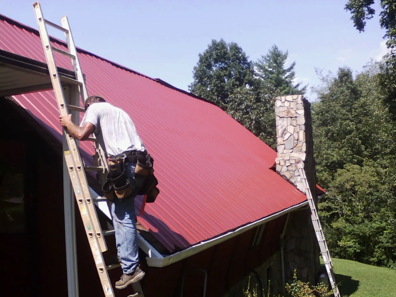 Roofer on a ladder beside a red metal roof and stone chimney — Soffit & Fascia Repair by Roofing Brothers in Calera